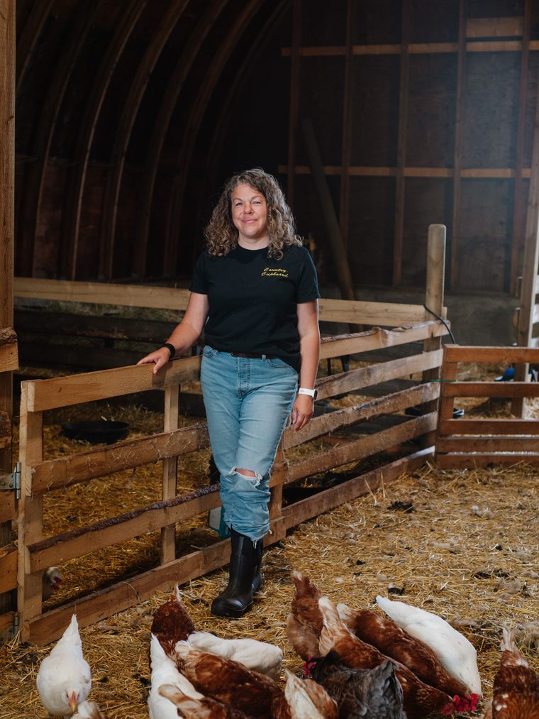 A farm shop owner standing in her barn with hens around her feet