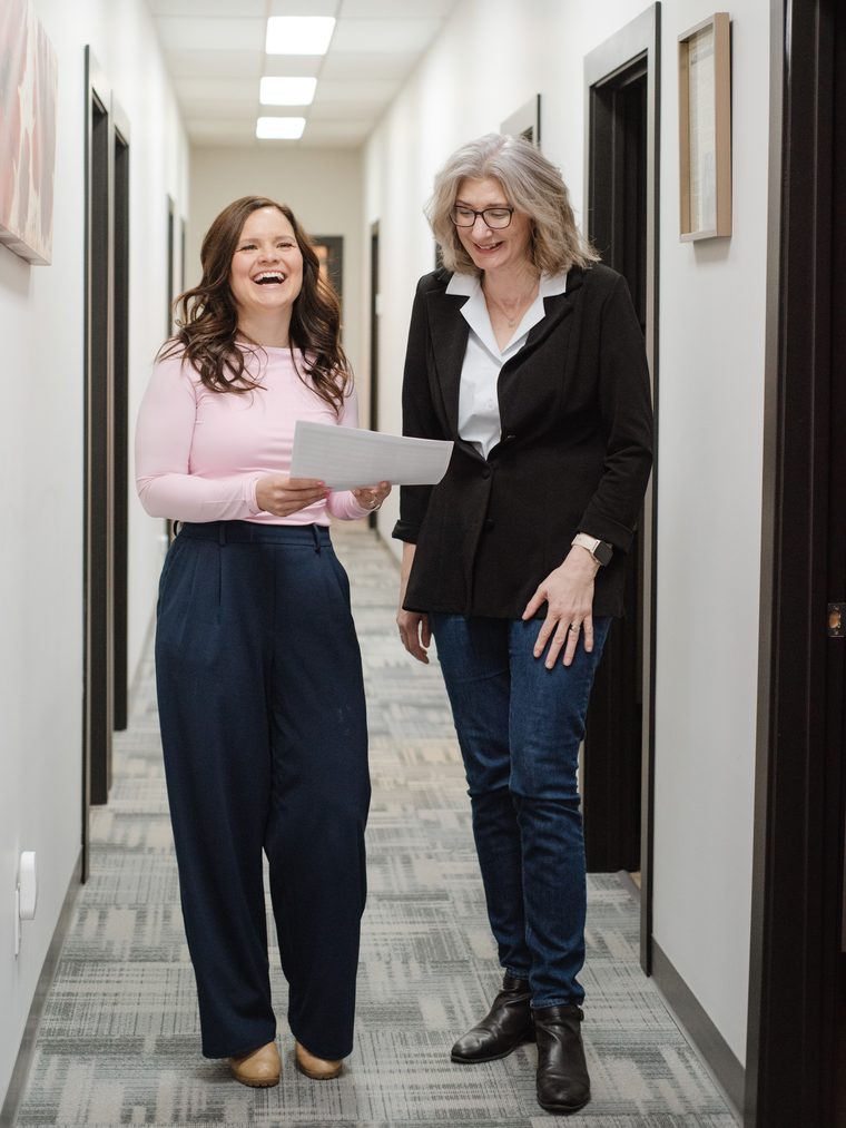 Two colleagues laughing together over a page in their office hallway